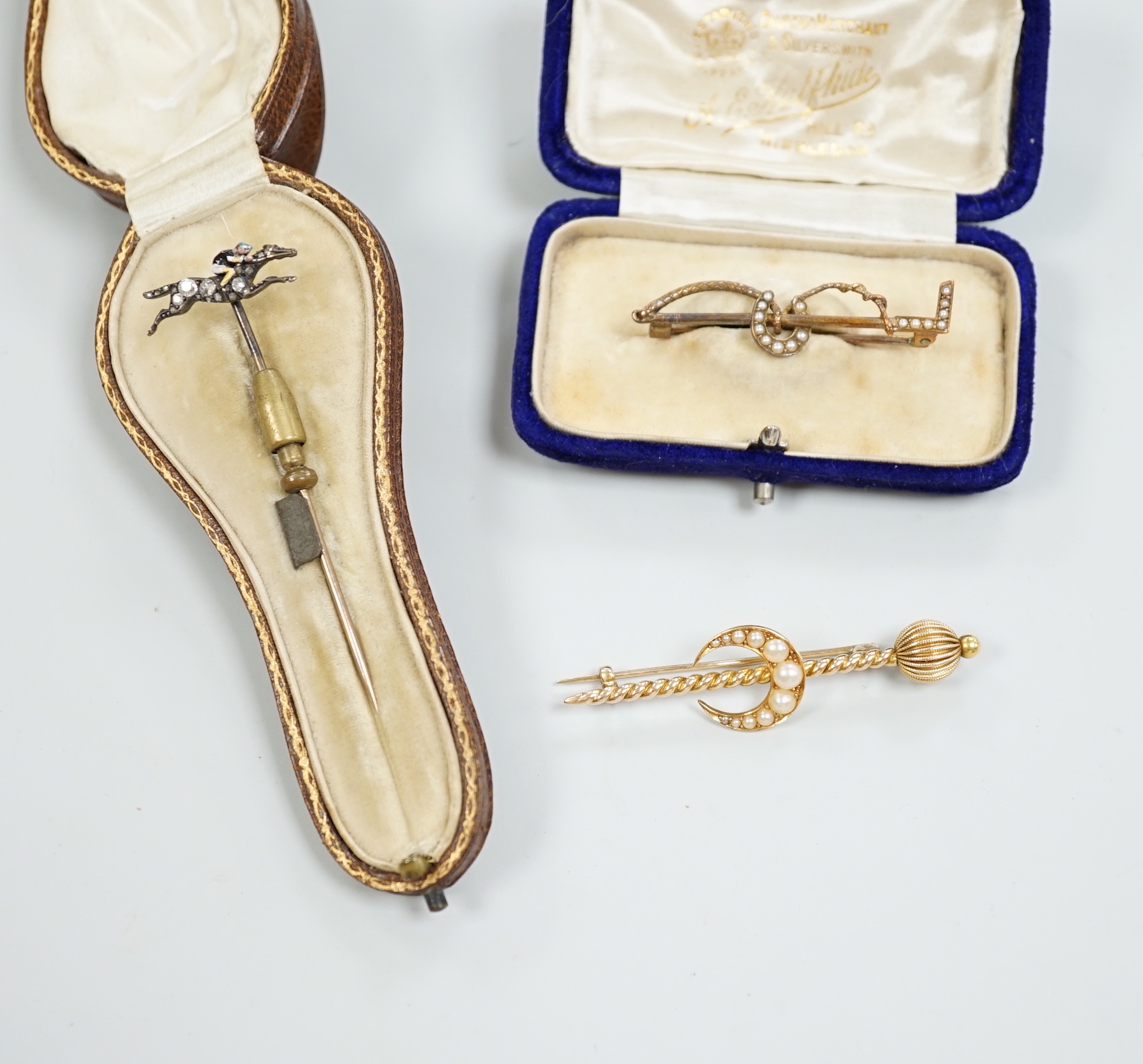 An Edwardian enamel and diamond set 'horse and jockey' stick pin, 63mm, a yellow metal and seed pearl set 'crescent and staff, brooch and a 'riding crop and horseshoe' brooch.